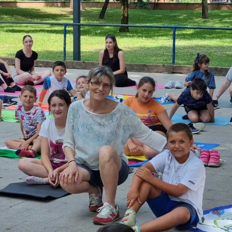 La dottoressa Barbara Camilli, psicologa a Novara, sorride accovacciata insieme a un gruppo di bambini seduti sui tappetini durante un laboratorio terapeutico ed educativo all'aperto nel verde.